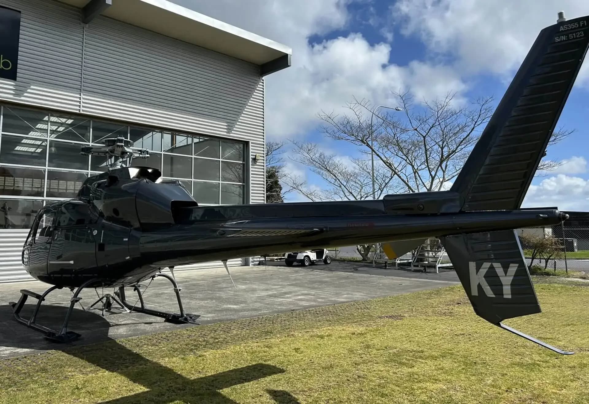 A rear-side view of a dark grey Eurocopter AS355 F1 helicopter, highlighting its tail boom with "KY" marking, parked next to a hangar.