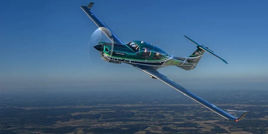 A green and black Diamond DA50 RG aircraft with 'DARK SHARK' livery flies against a blue sky over a forested landscape.