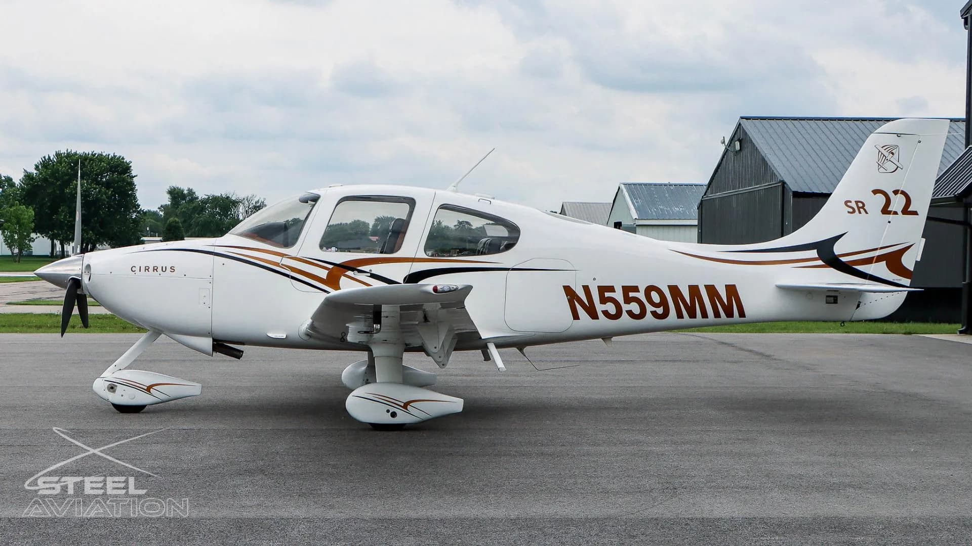 A white Cirrus SR22 aircraft with N559MM registration is parked on an airfield with trees and buildings in the distance.