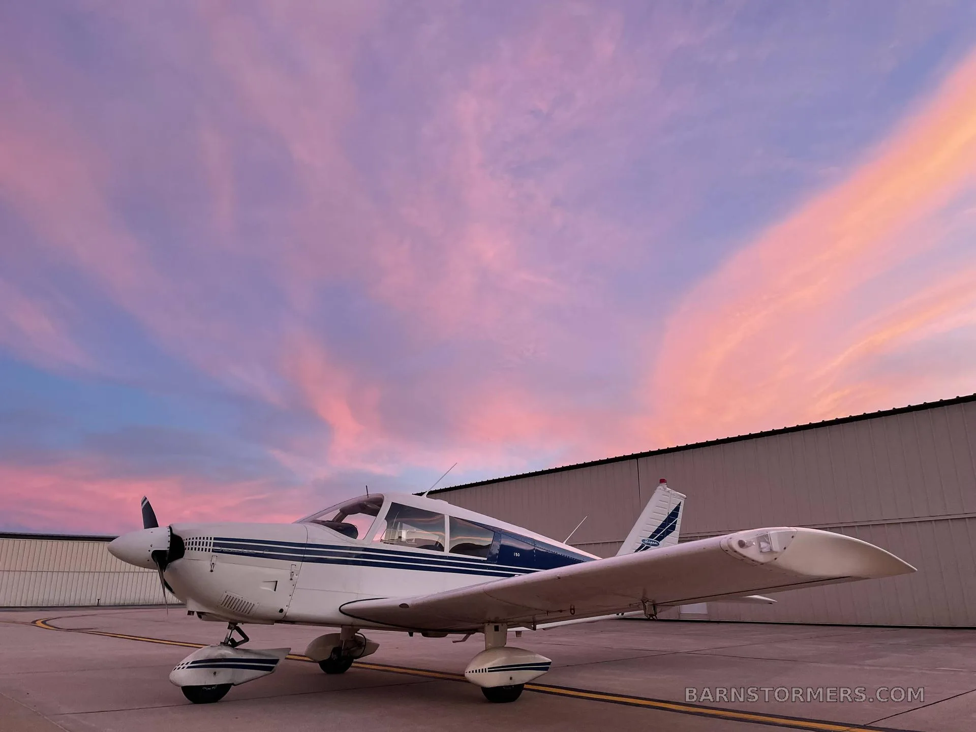 A white Piper Cherokee 150 aircraft with blue stripes parked on an tarmac at sunset, with a hangar in the background.