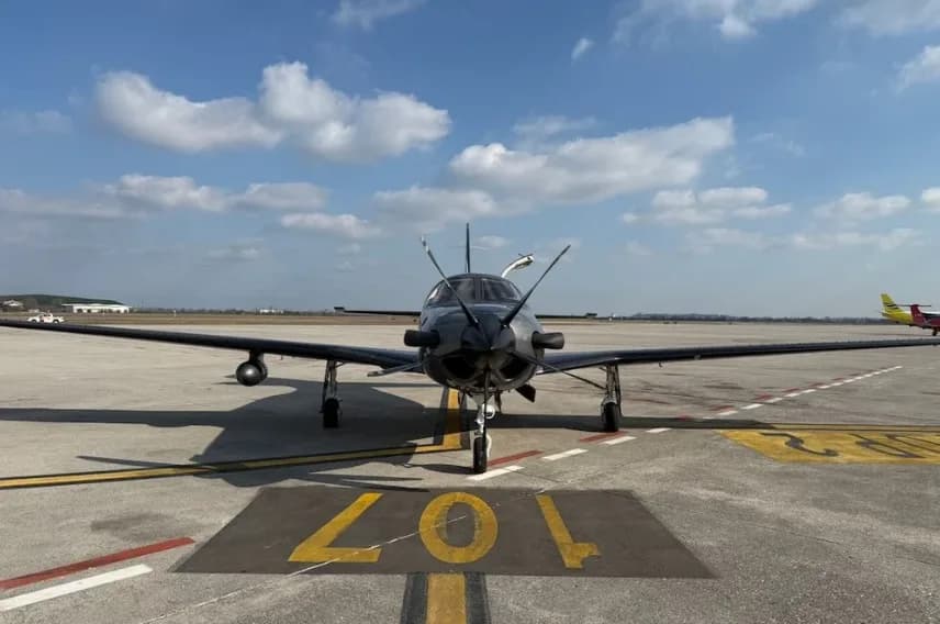 A dark grey Piper M600 SLS aircraft is parked on an airfield with runway markings, viewed from the front.