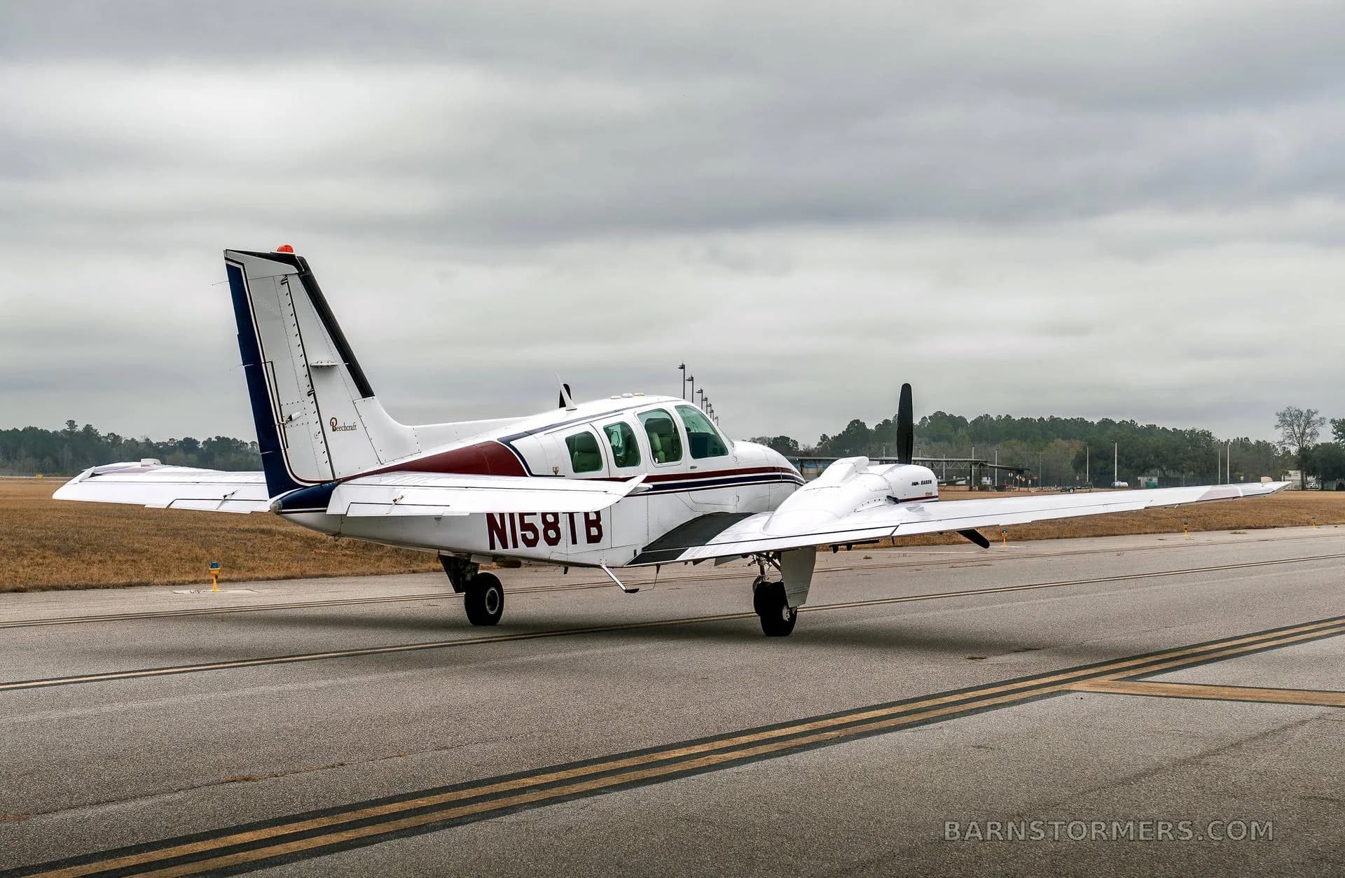 A white twin-engine Beechcraft Baron N158TB with maroon and blue stripes is seen from the rear-right on a taxiway under a cloudy sky.