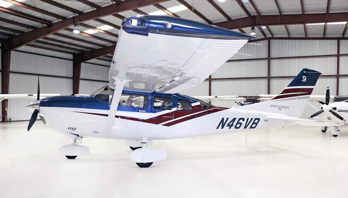 A white and blue Cessna Stationair TC aircraft (N46VB) with maroon accents is parked in a hangar, viewed from the front-right.
