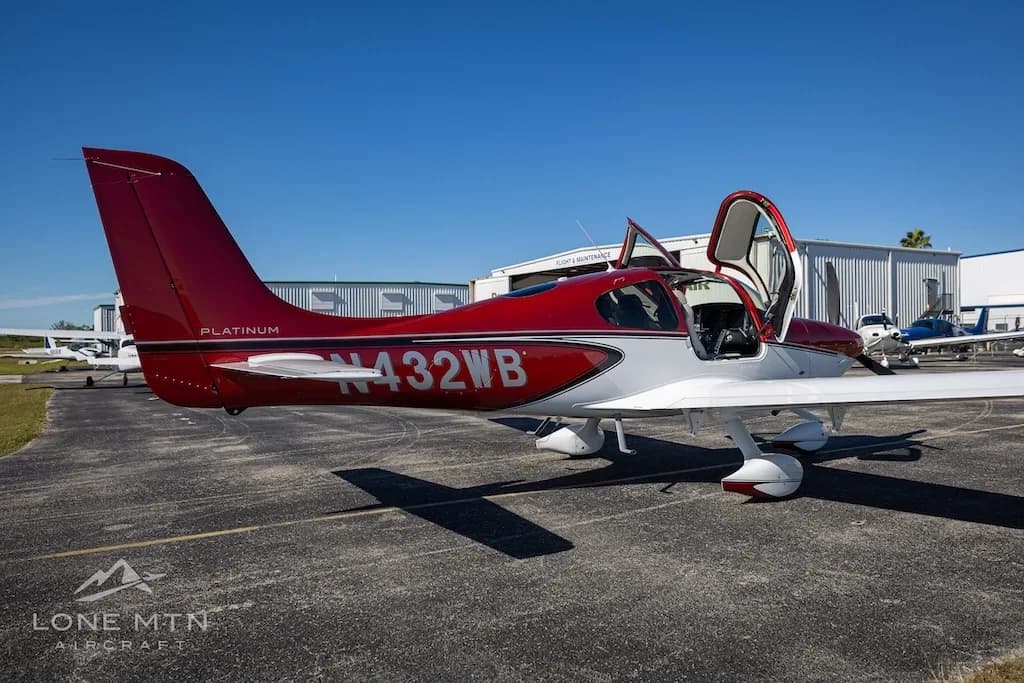 A rear-side view of the red and white Cirrus SR20 aircraft (N432WB) with its doors open, parked near a hangar.