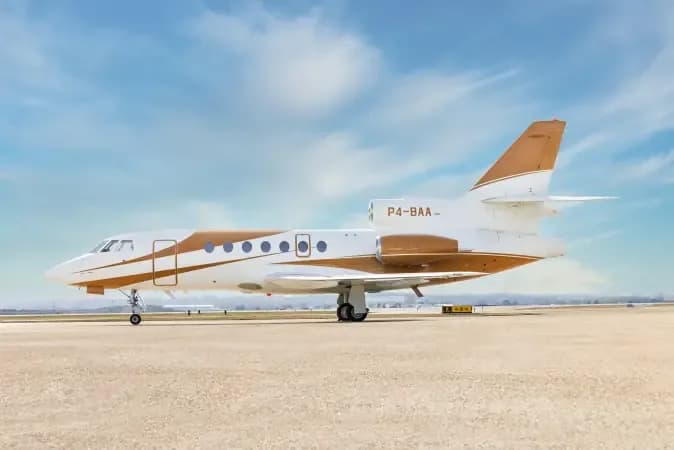 A side view of the white and gold Dassault Falcon 900EX business jet with registration P4-BAA parked on an airfield.