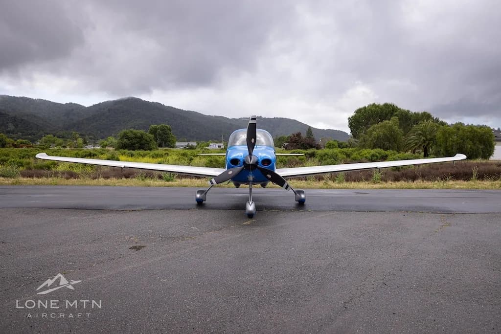 A front view of a blue and silver single-engine propeller aircraft (Cirrus SR22) with registration N248LF parked on a wet tarmac under a cloudy sky.