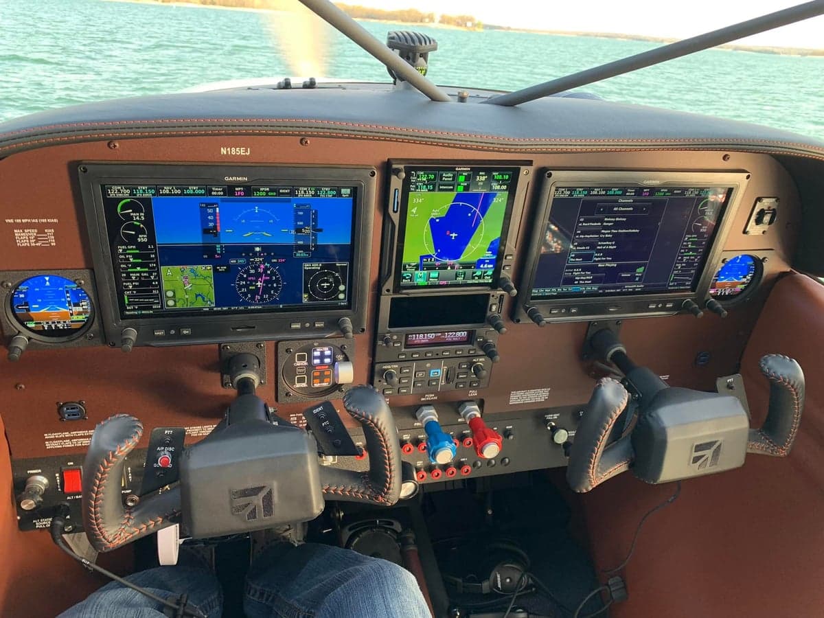 View of the Cessna 185 instrument panel with modern avionics, with a lake visible outside.