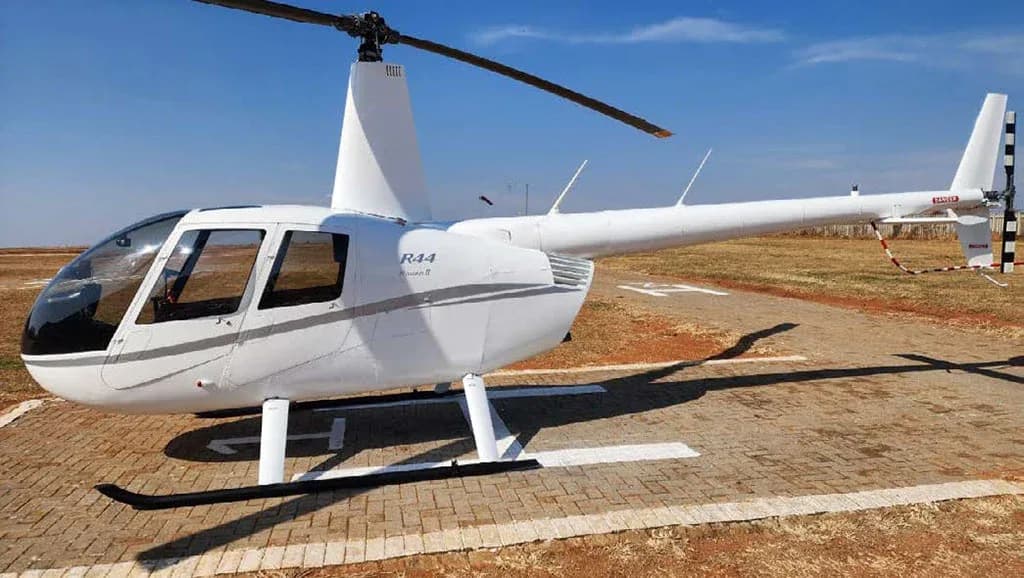 A white Robinson R44 Raven II helicopter is parked on a paved surface with a grassy field in the background.