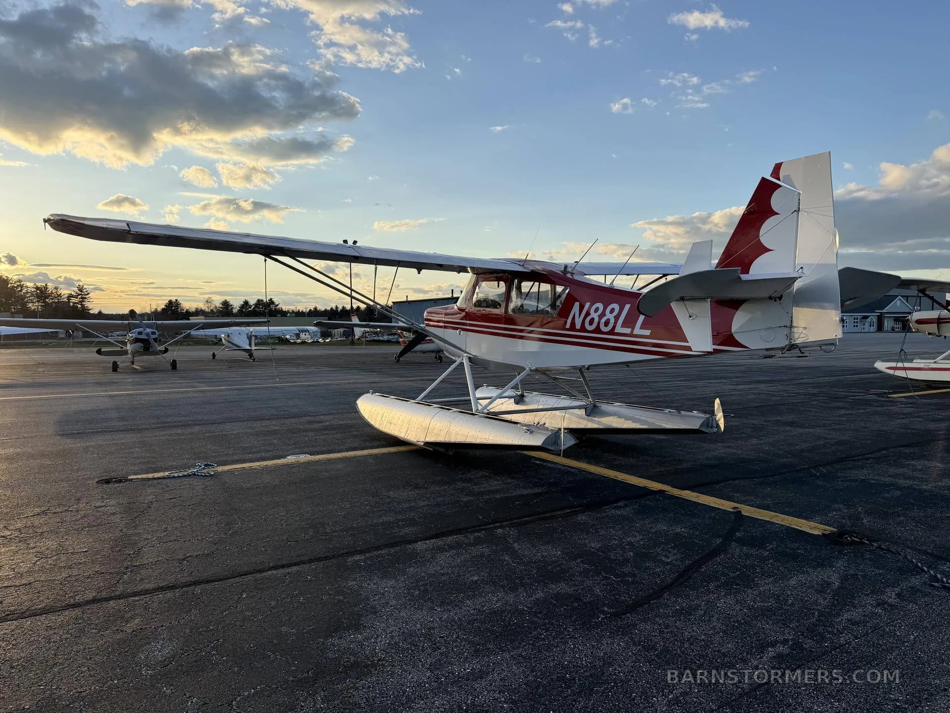 A red and white floatplane with tail number N88LL, parked on an asphalt runway at sunset with other aircraft in the background.