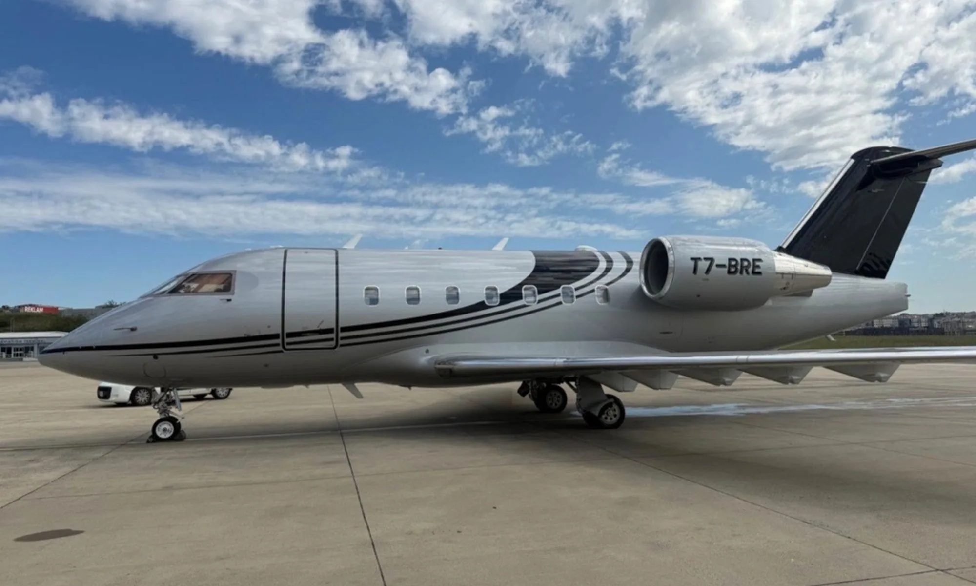 A silver private jet with black stripe livery, registration T7-BRE, parked on an tarmac under a blue sky.