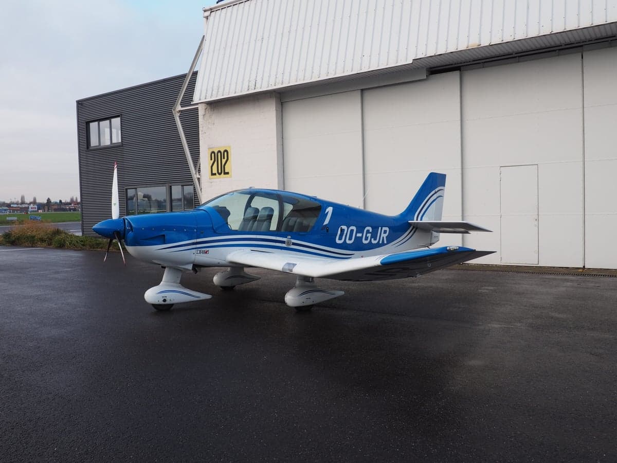 A blue and white light aircraft, registration OO-GJR, parked outside a hangar.