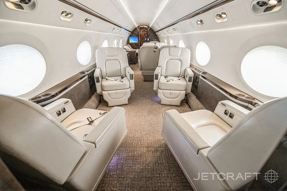 A view down the aisle of a private jet cabin, showing pairs of light-colored leather seats facing each other.