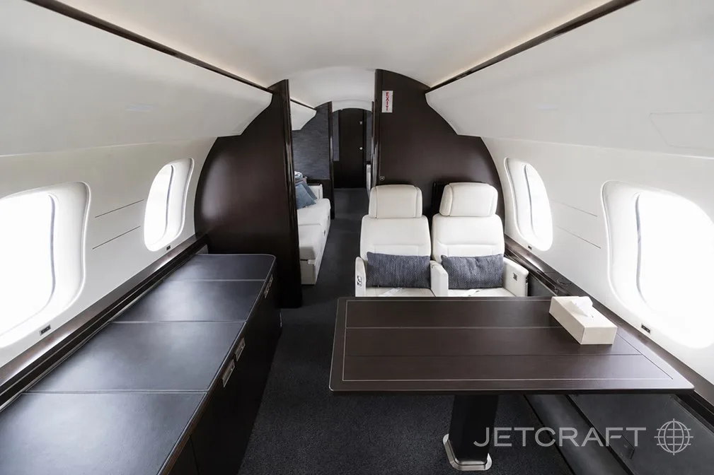 A private jet cabin with a dark wood table, white seats, and a lounge area visible further back.