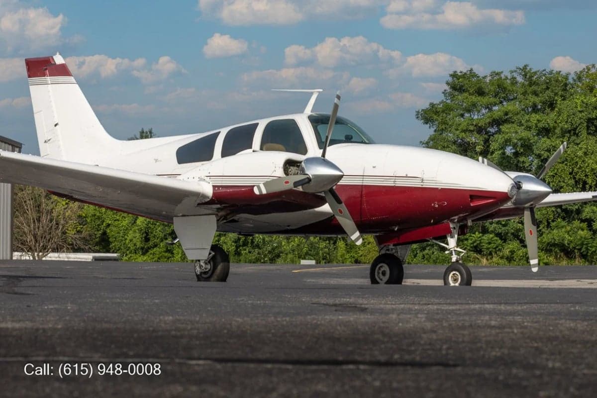 A white and red twin-engine propeller aircraft, likely a Beechcraft Baron, parked on an asphalt ramp with trees and a cloudy sky.