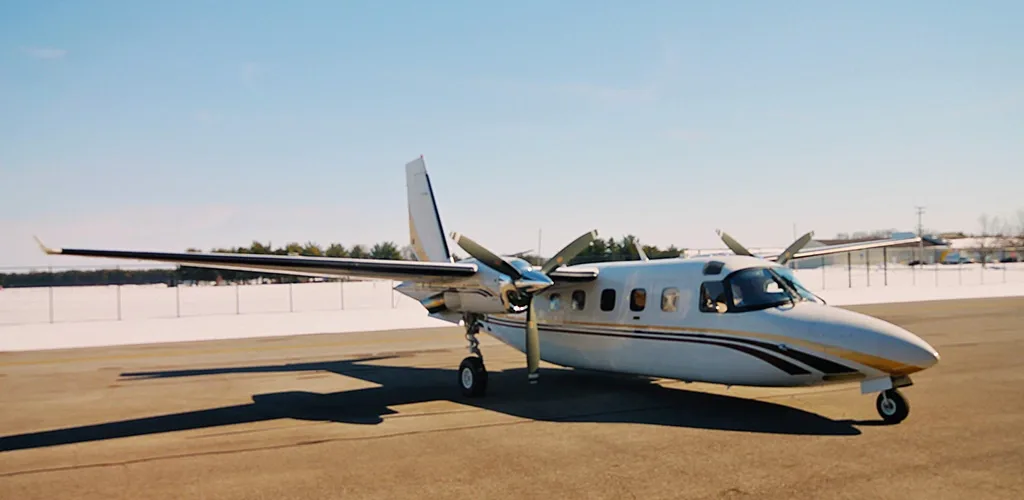 A white twin-engine turboprop aircraft with gold and maroon stripes parked on an airfield with snow-covered ground in the distance.
