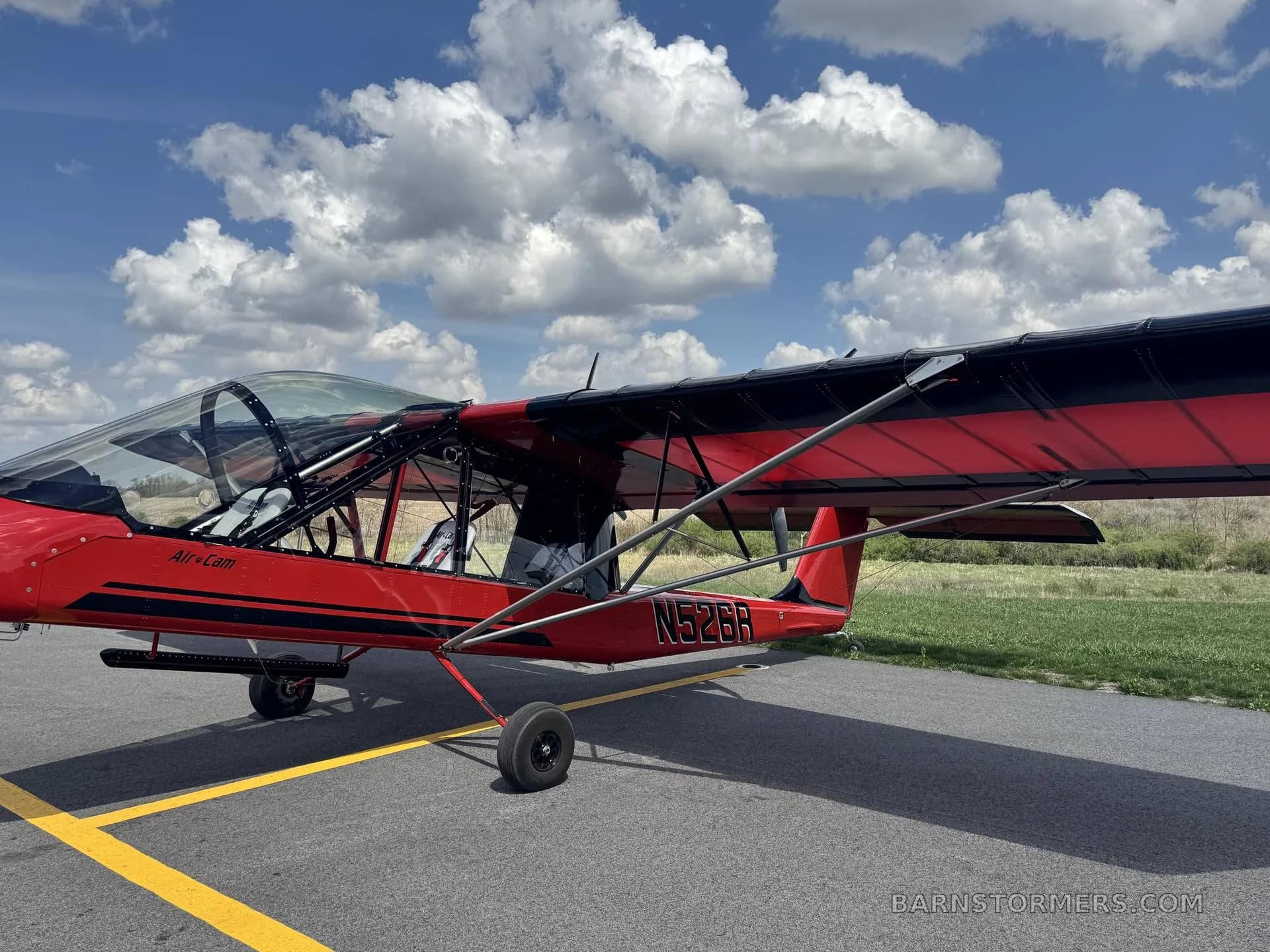 A red and black Air-Cam aircraft with registration N526R is parked on an asphalt runway with green fields in the background.