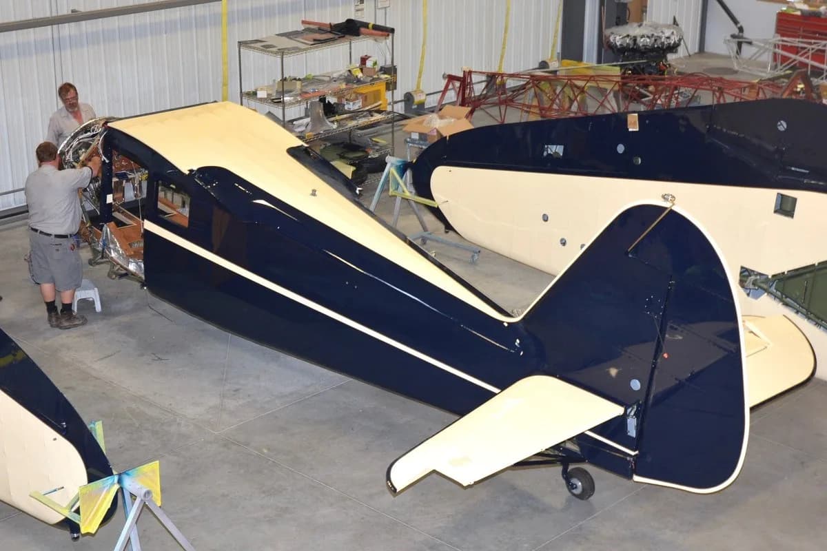 Two men are working on the dark blue and cream fuselage of a vintage aircraft inside a hangar.
