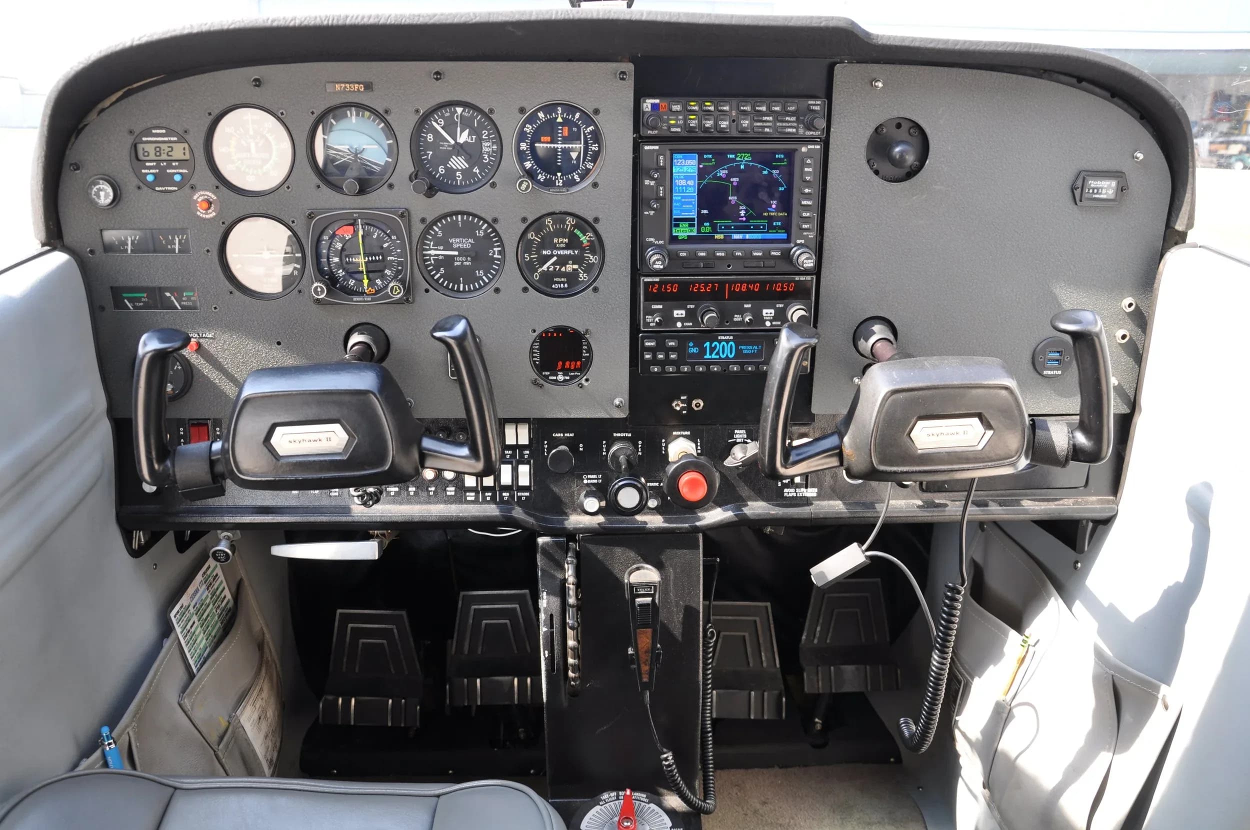 The cockpit of a Cessna aircraft, showing the instrument panel with analog gauges and a modern GPS display, yokes, and rudder pedals.