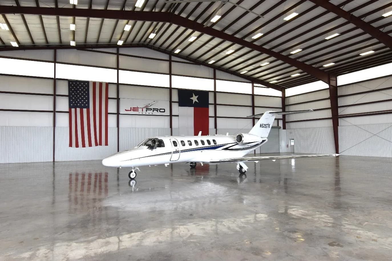 A white Cessna Citation CJ3 private jet with blue and black stripes, registration N525TK, parked in a hangar with US and Texas flags.