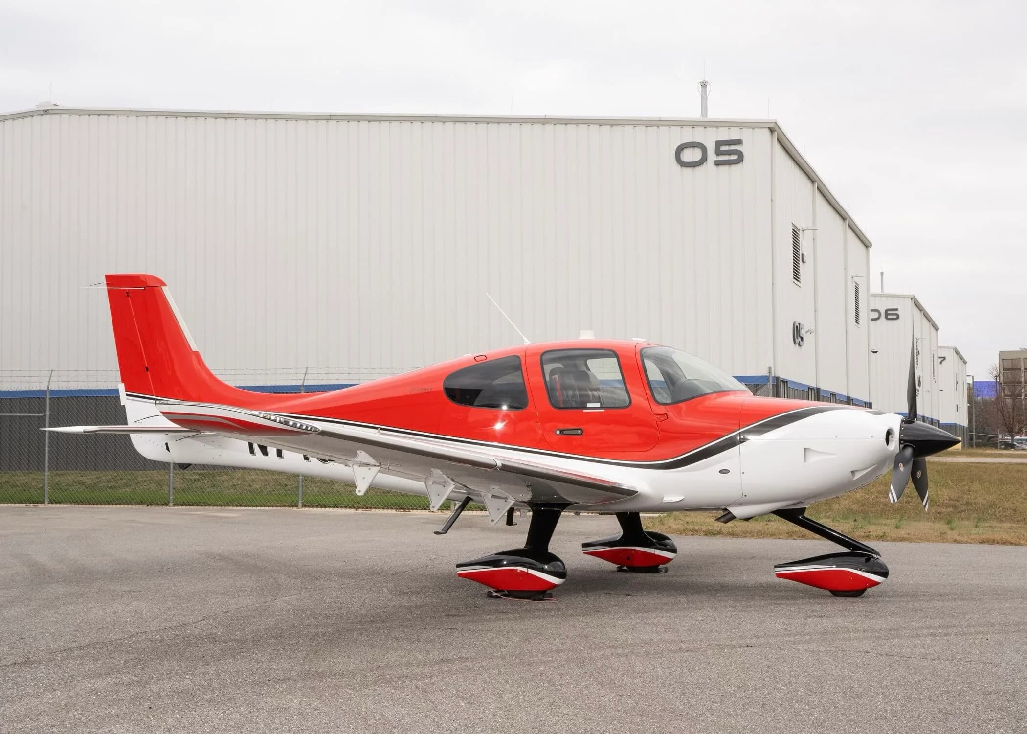 A red and white Cirrus SR22 aircraft with registration N715CH parked on an airfield in front of hangars.