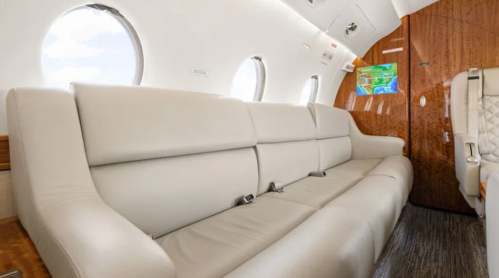 A close-up view of the long beige leather sofa inside a private jet cabin, with windows visible.