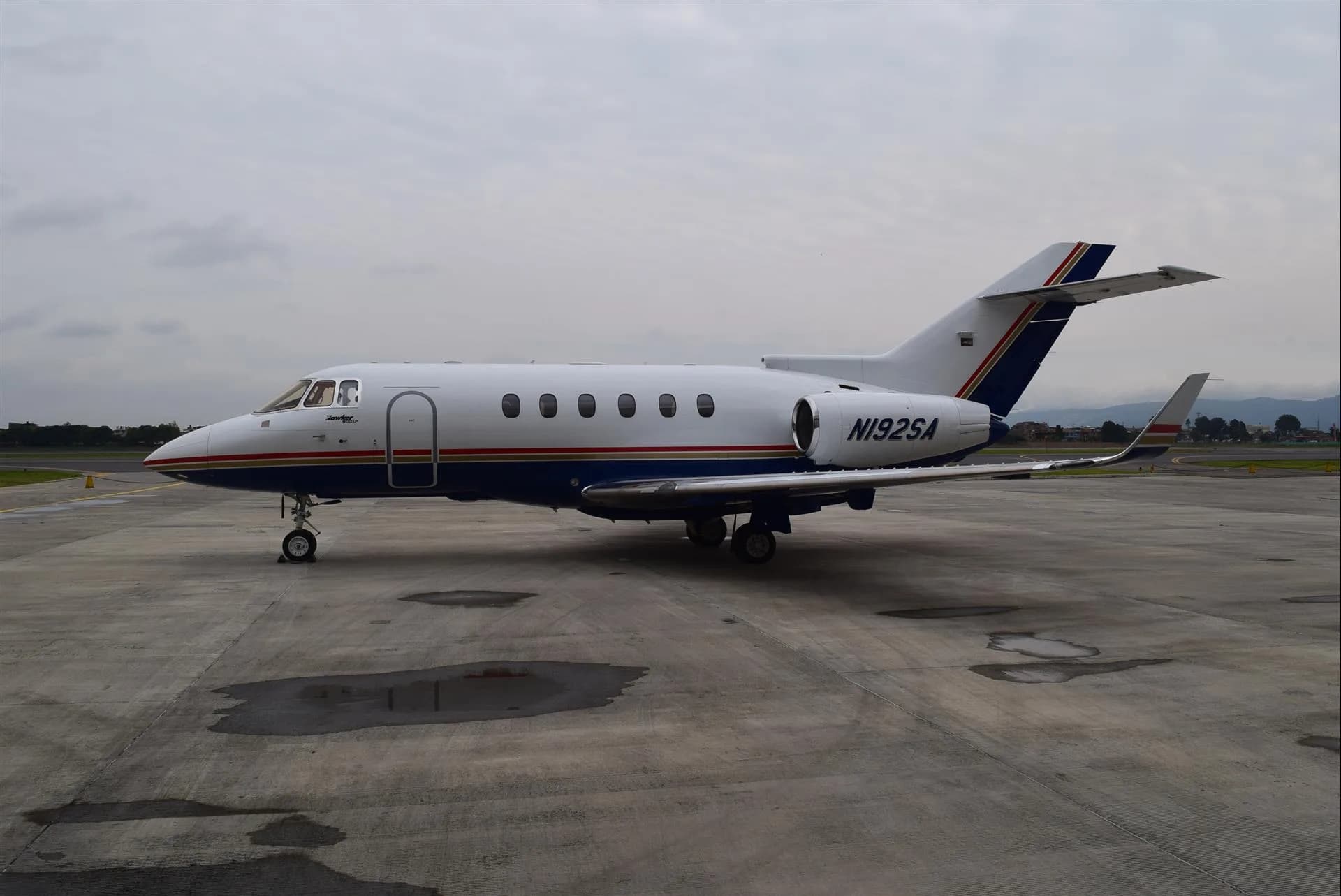 Side view of a white and blue Hawker jet with tail stripes on the tarmac.