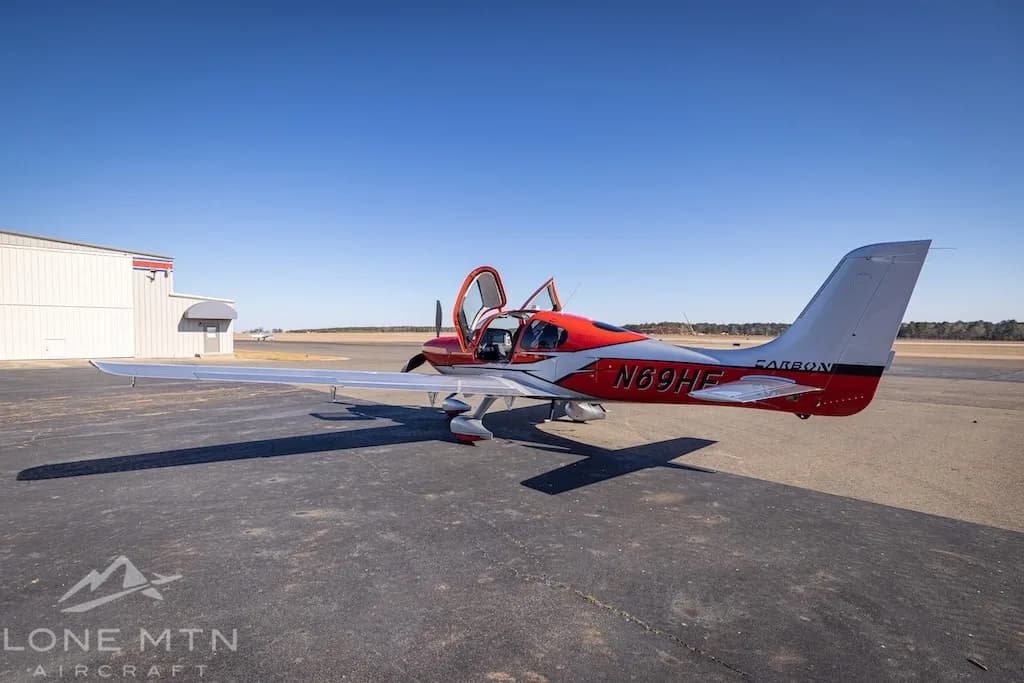 A red and silver Cirrus SR22T aircraft with open doors parked on an asphalt ramp next to a hangar.