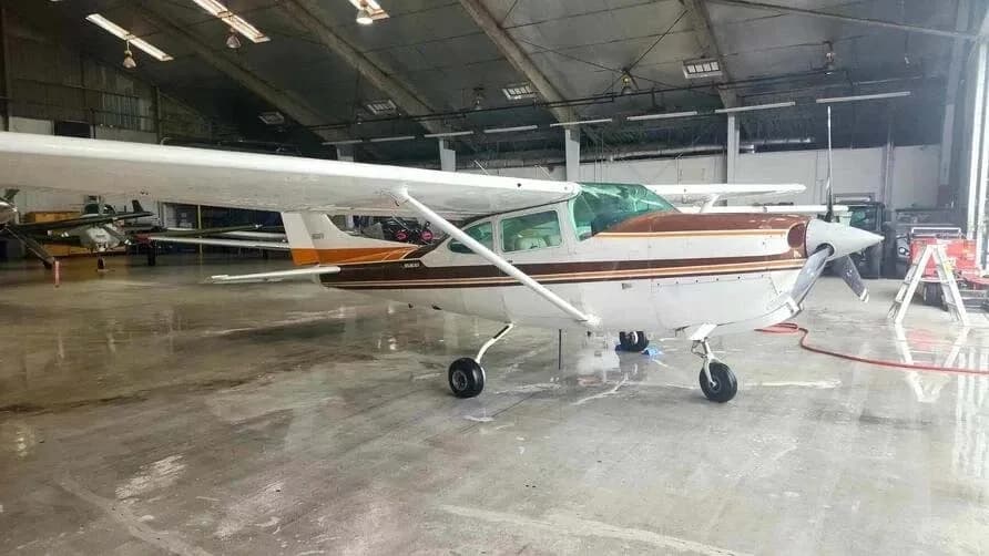 A white and brown Cessna Skylane RG II aircraft parked inside a large hangar with other aircraft in the background.