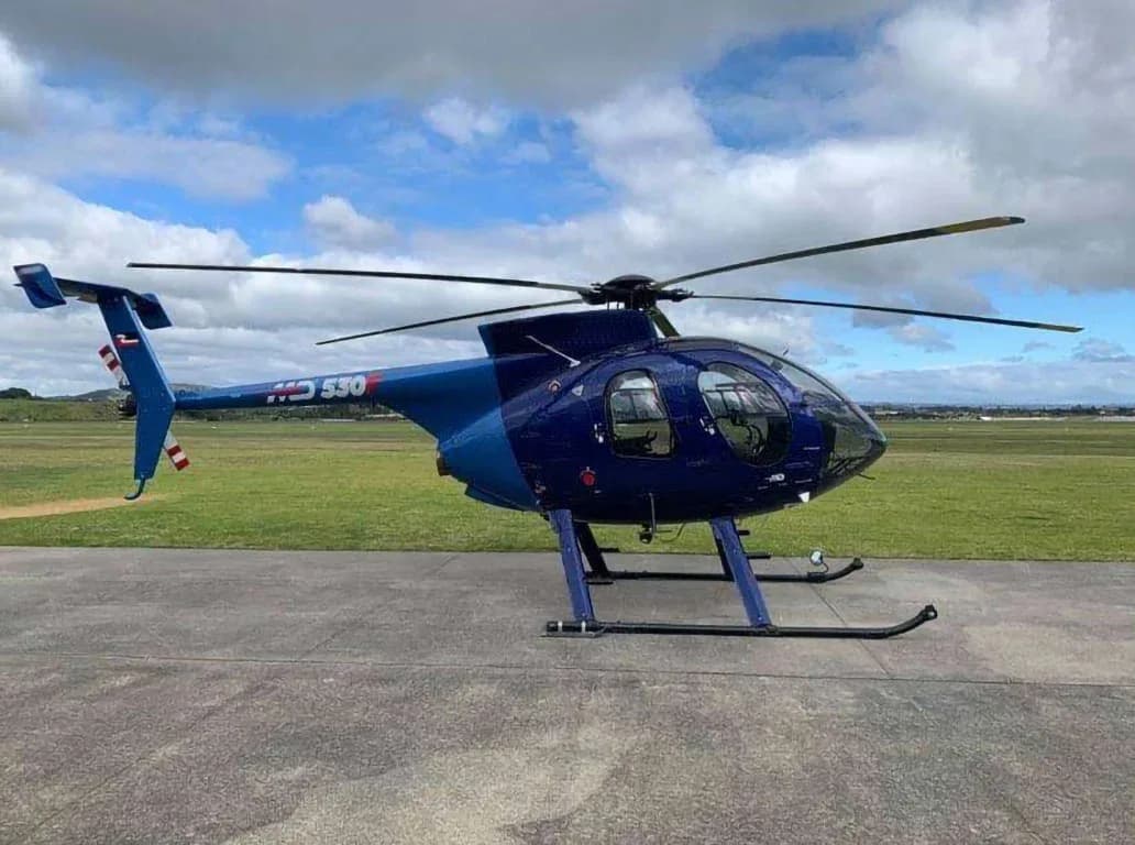 A blue and light blue MD 530 helicopter with 'MD 530' on its tail, parked on an airfield under a cloudy sky.