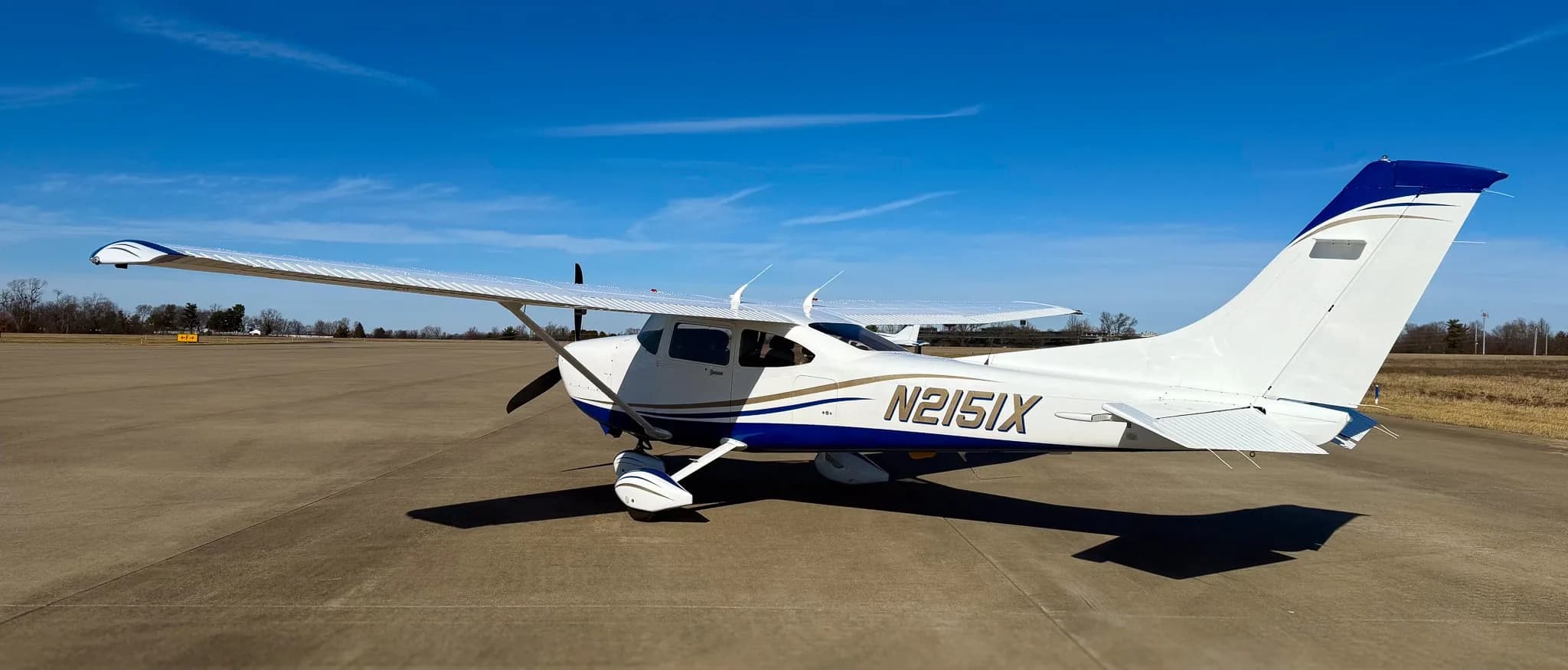 A white and blue Cessna 182 Skylane (N2151X) parked on a tarmac, viewed from the rear-right.