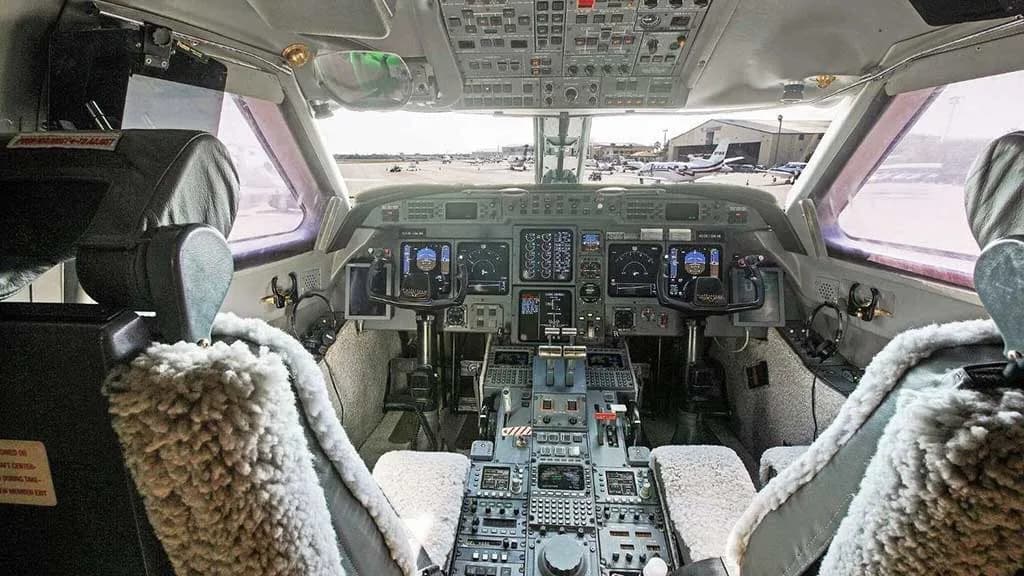 The cockpit of a Gulfstream G550 aircraft, showing the instrument panel and controls with a view of the airfield outside.