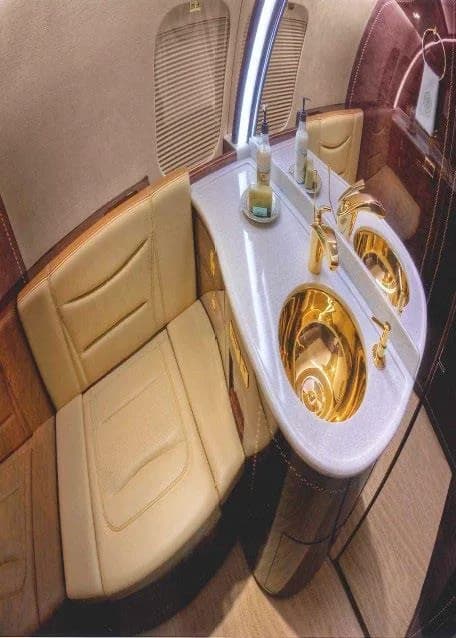 A private jet lavatory featuring a golden sink, a mirror, and a beige leather seat.