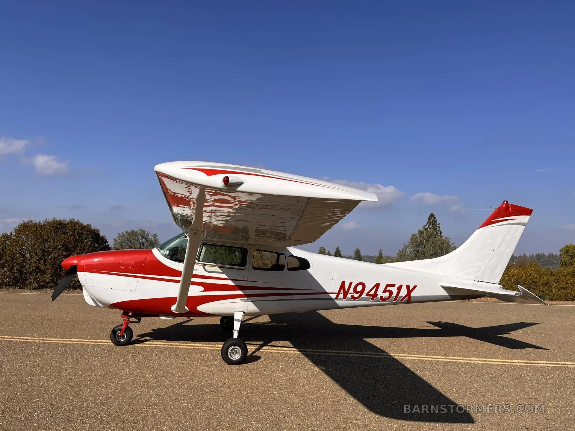 A white and red Cessna 182 aircraft with registration N9451X parked on an asphalt surface under a clear blue sky.
