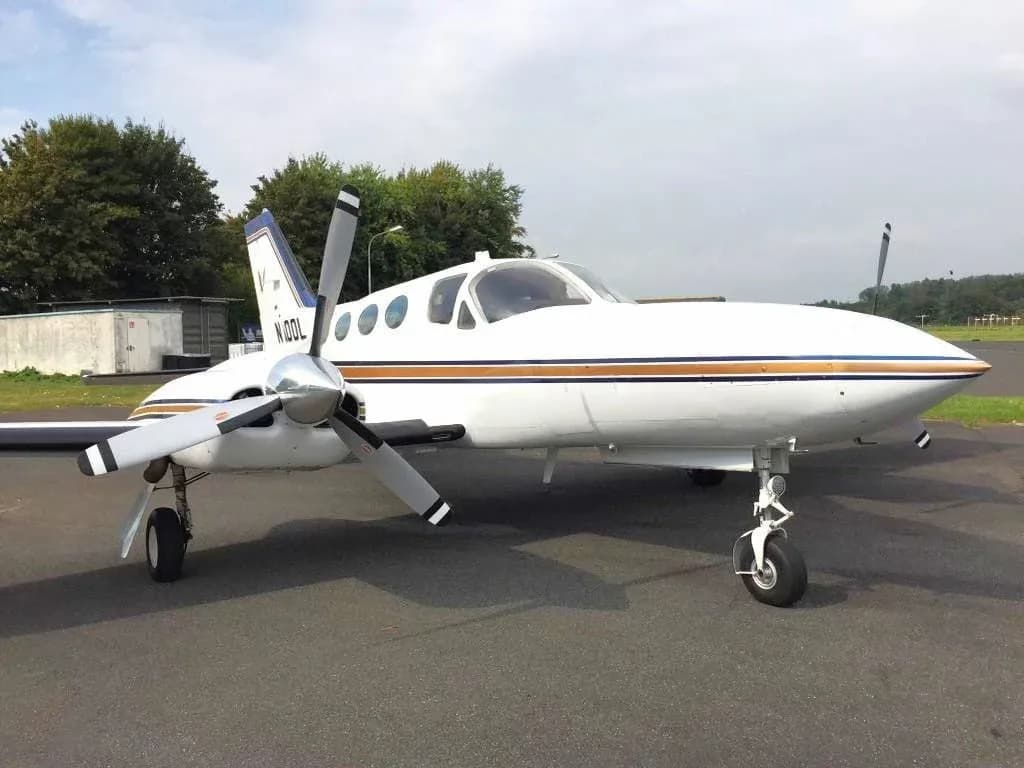 A white Cessna 421 Golden Eagle aircraft with blue and gold stripes parked on the tarmac with trees in the background.