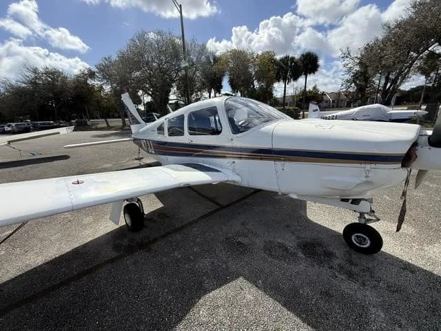 A front-side view of the white Piper Cherokee aircraft, showing its propeller and blue and red stripes.