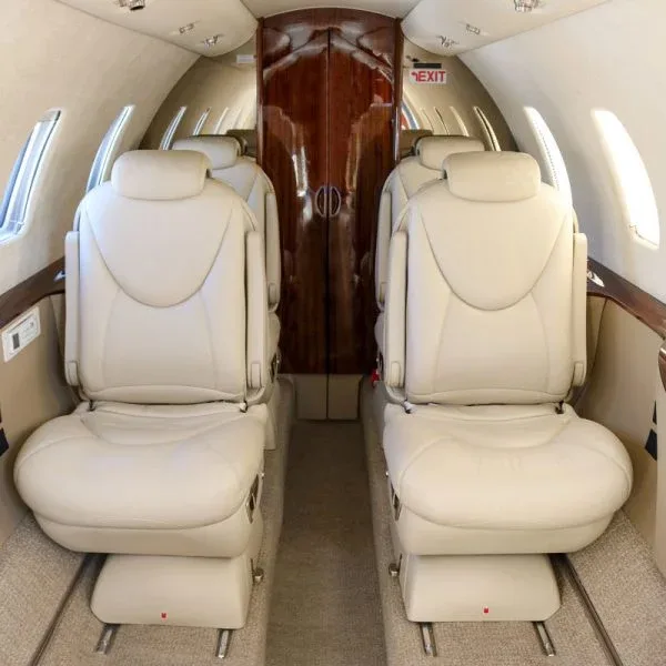 View down the aisle of a private jet cabin with light beige leather seats and dark wood accents.