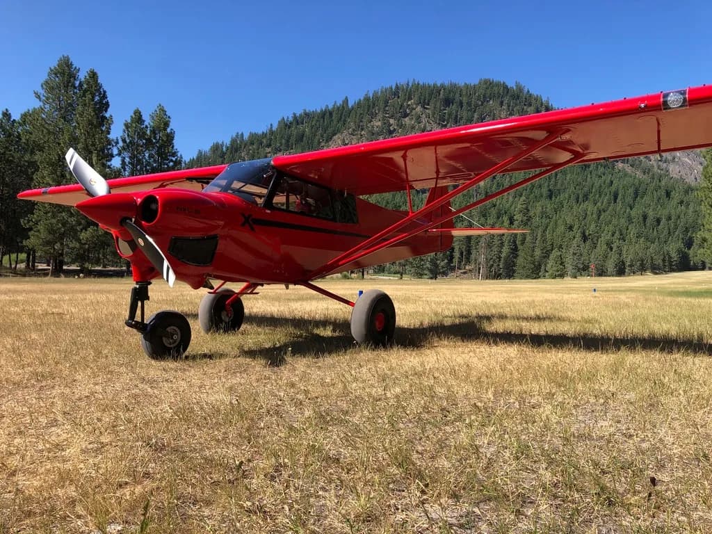A red CubCrafters XCub parked on a grassy field with trees in the background.