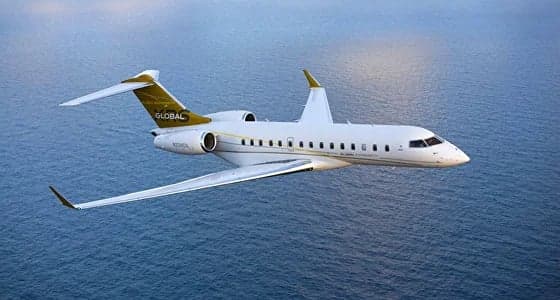 A white Bombardier Global business jet with gold tail accents flies over a blue ocean.