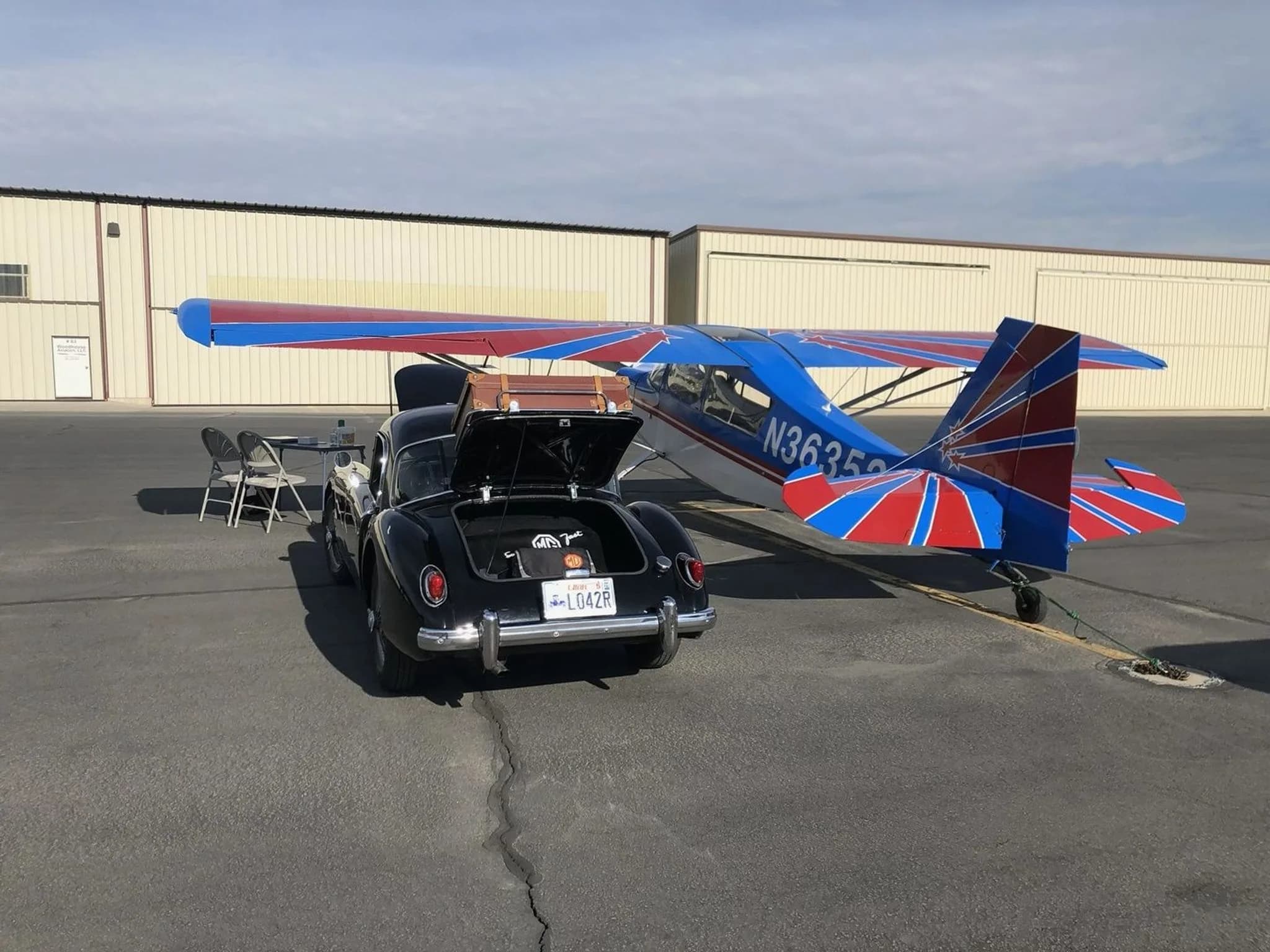 A blue and white high-wing aircraft, N36352, parked on an airfield tarmac next to a black classic car.