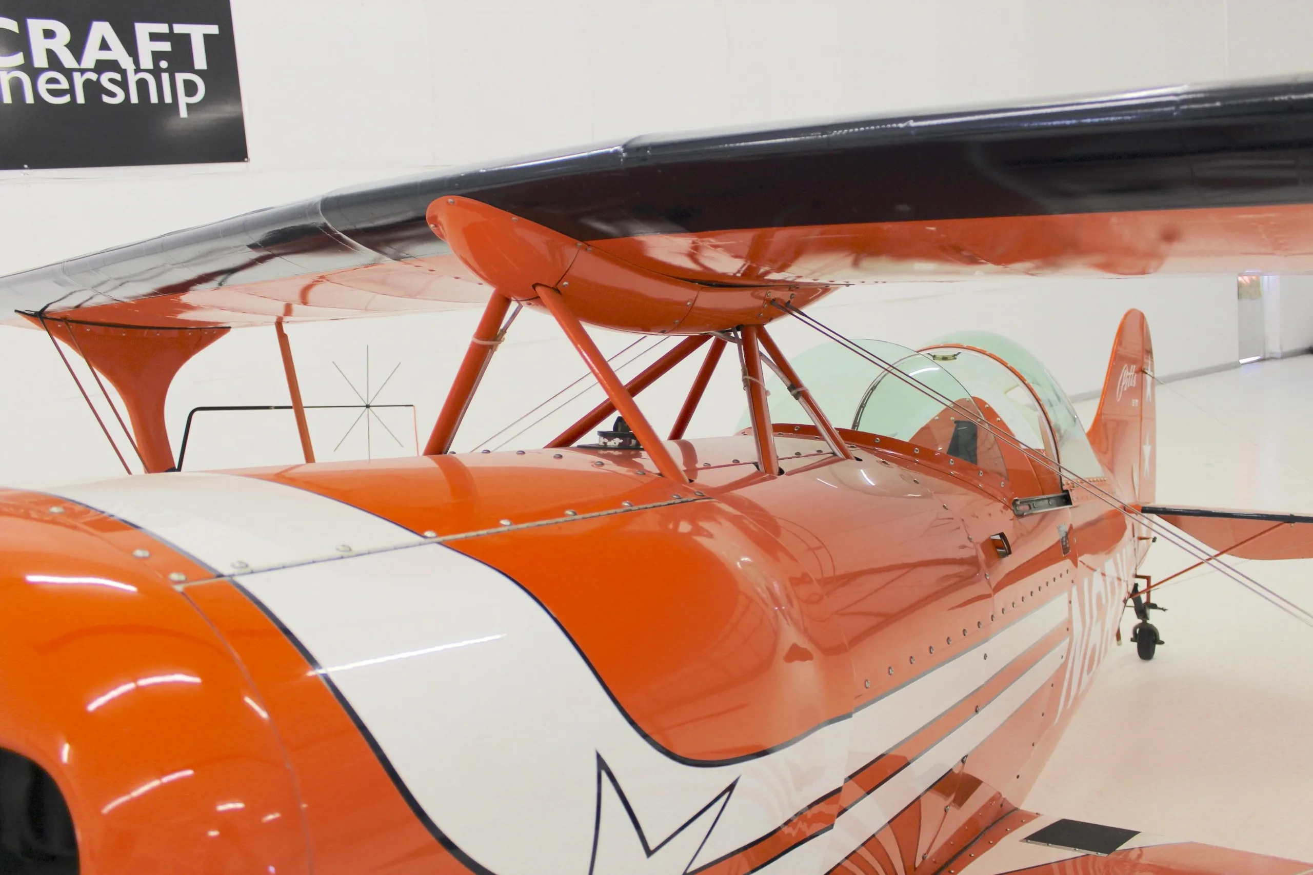 A close-up view of the cockpit area and upper wing of an orange Pitts S-1T biplane.