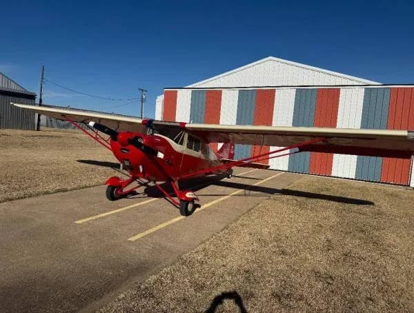 A red and cream high-wing taildragger aircraft parked on an airfield next to a striped hangar.