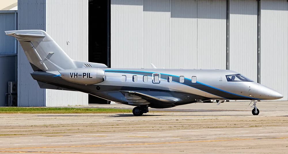 Side view of a silver and blue Embraer Phenom 300 with registration VH-PIL parked in front of a hangar.