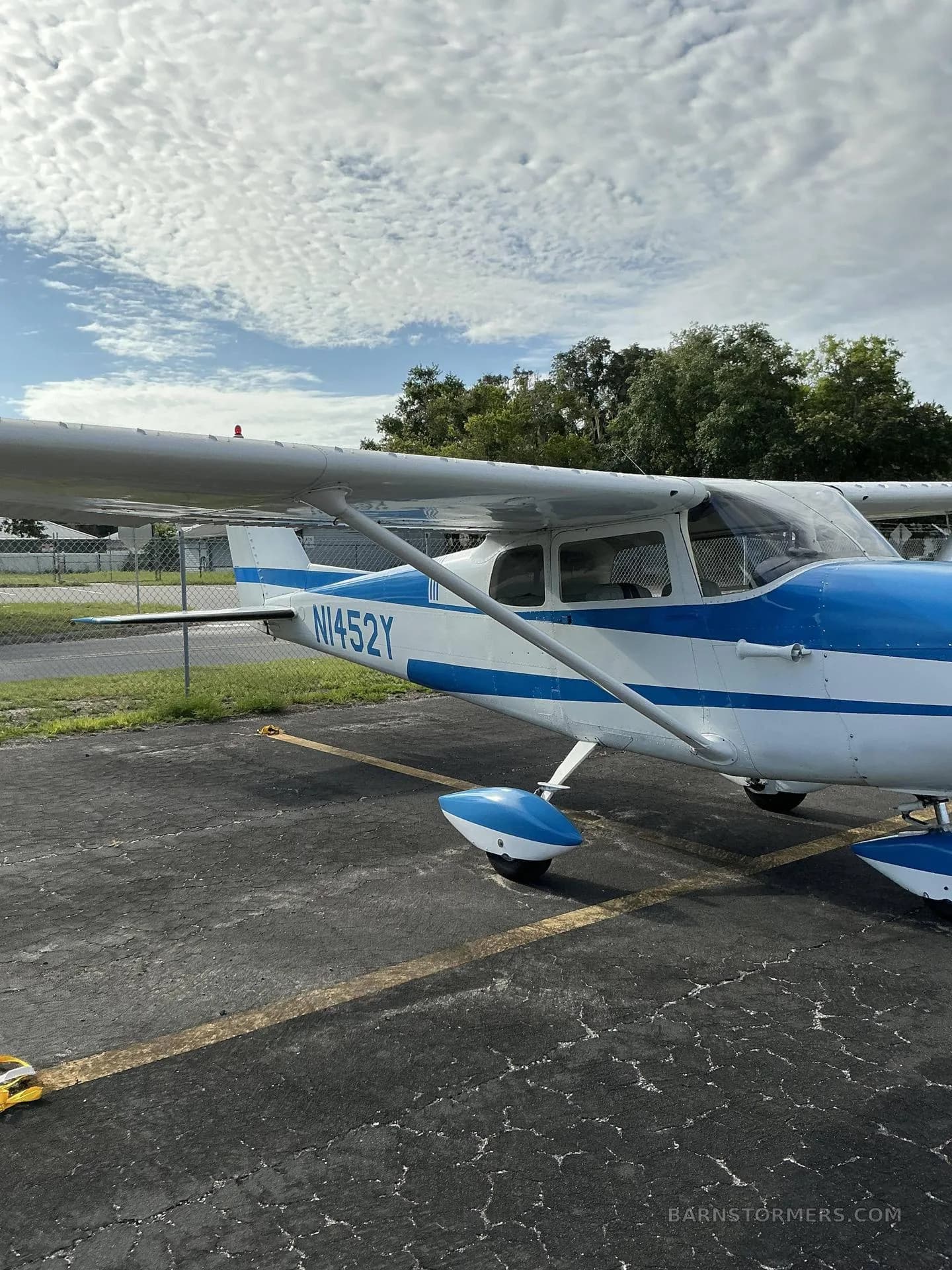 A white and blue Cessna 172 aircraft with tail number N1452Y is parked on an asphalt ramp next to a chain-link fence, viewed from the right side.
