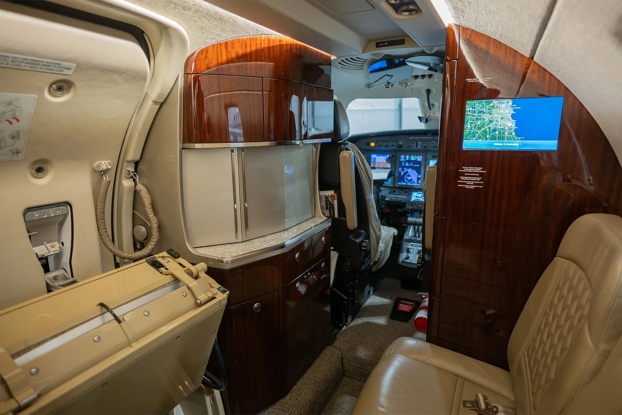 The forward cabin area of a business jet, featuring a galley, wood cabinetry, and a screen displaying a map, with the cockpit visible.