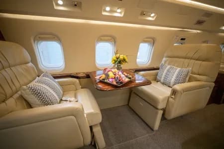 A luxurious aircraft cabin interior with two beige leather seats facing each other, a wooden table with fruit and flowers, and windows.