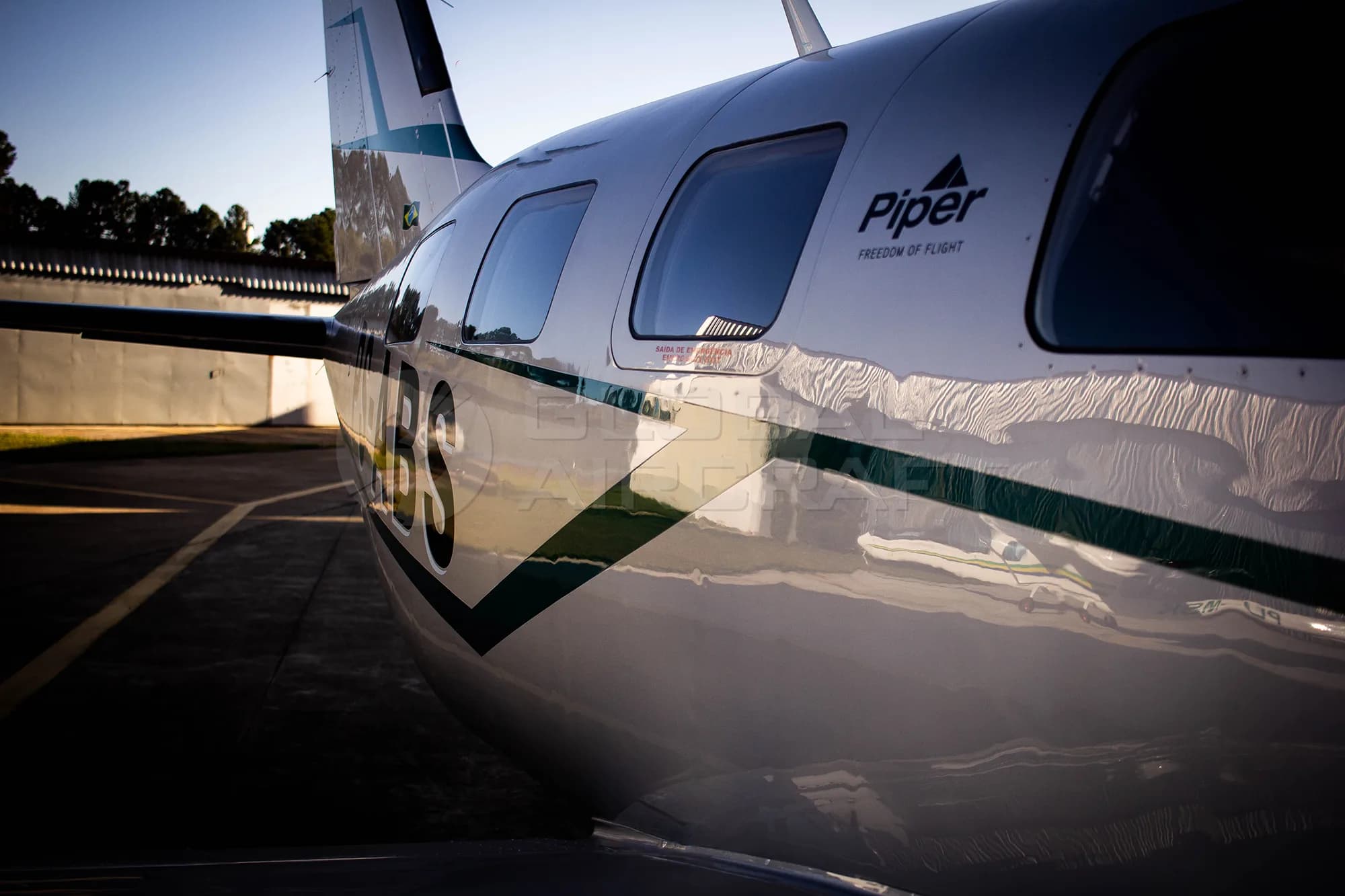 A close-up side view of the fuselage of a silver Piper M350 aircraft, showing the tail number PS-ABS and the Piper logo.