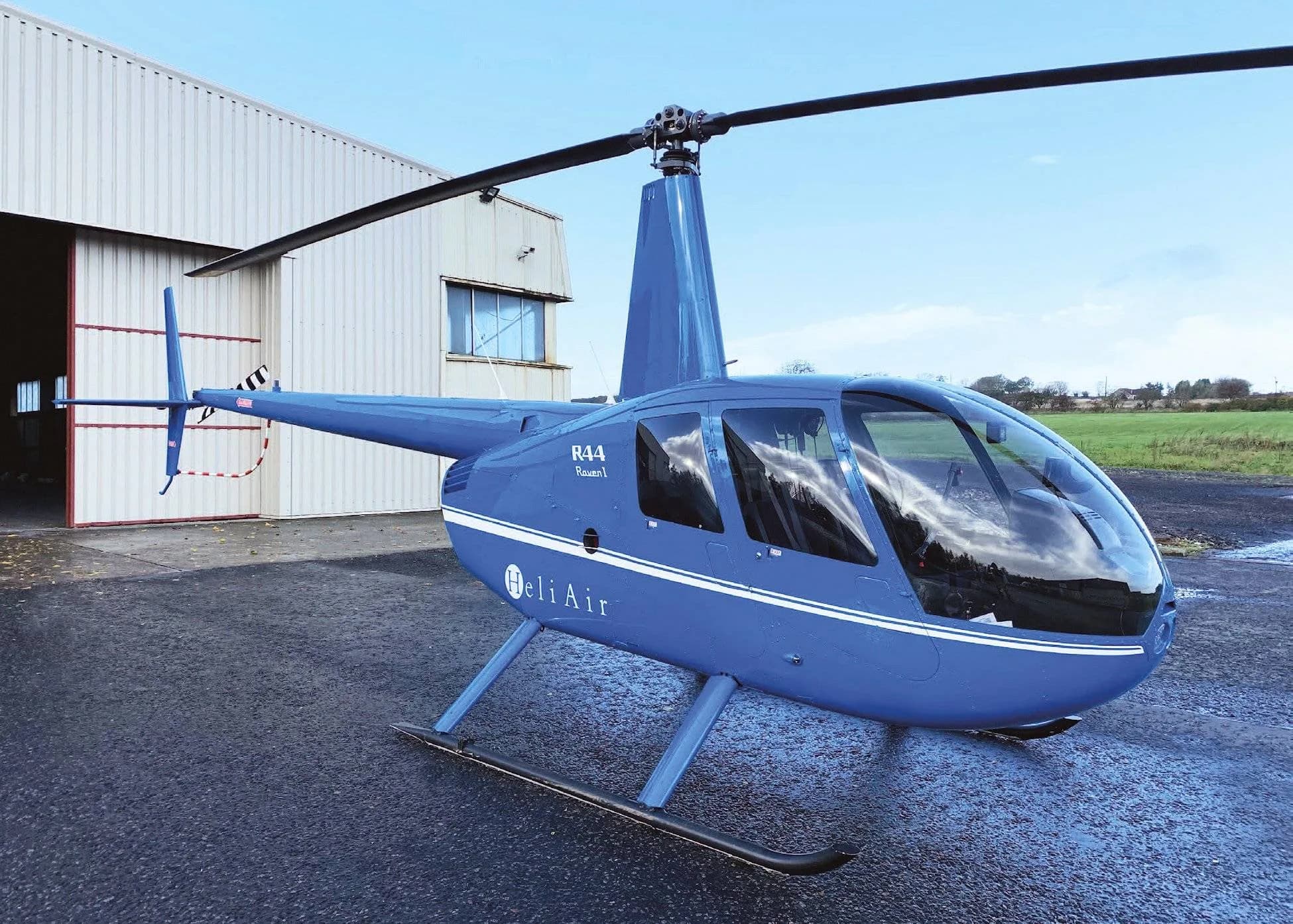 A blue Robinson R44 Raven II helicopter with a 'Heli Air' logo is parked on a wet tarmac in front of a hangar.