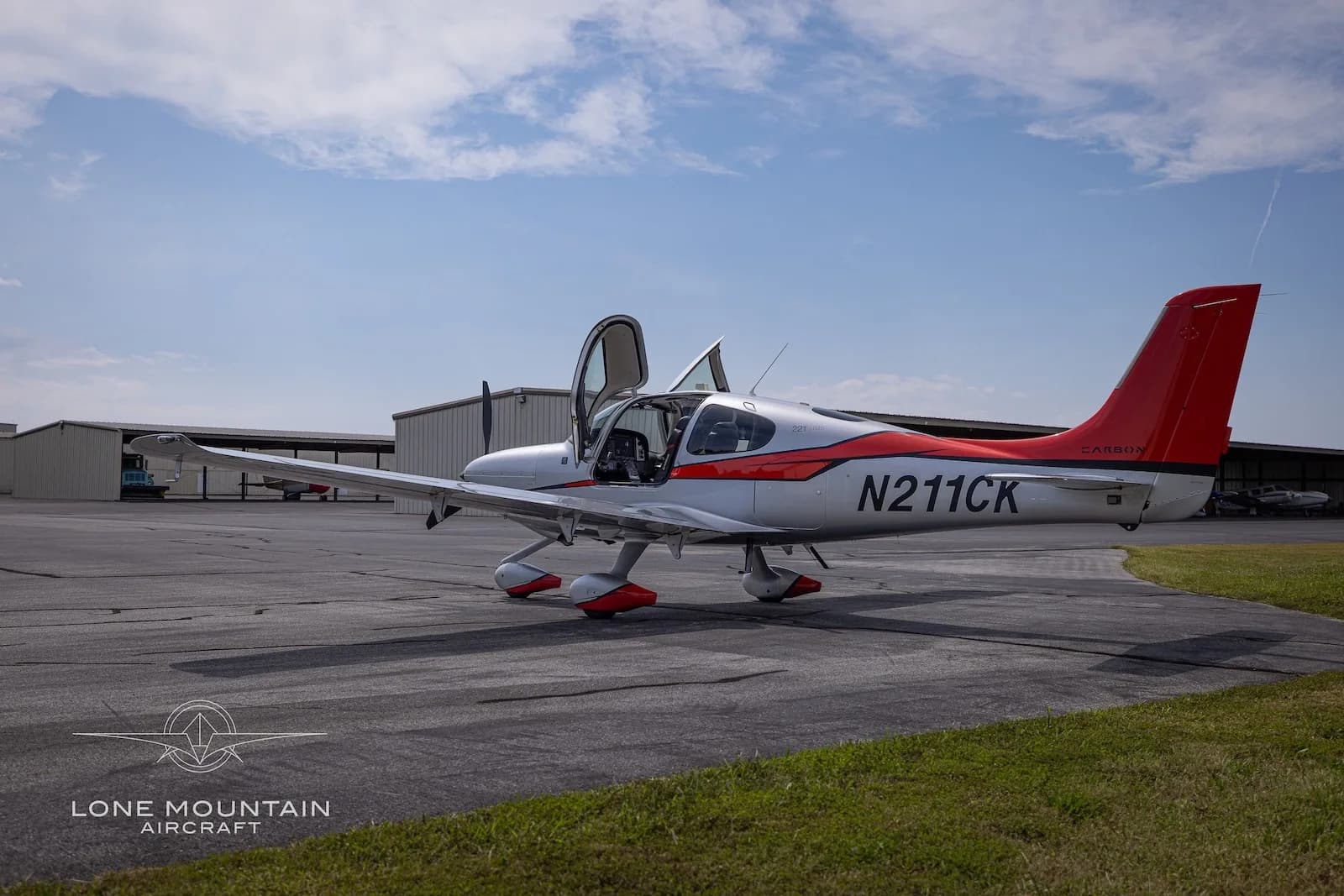 A three-quarter side view of a silver and red Cirrus SR22T aircraft with registration N211CK parked on an asphalt ramp with its gull-wing doors open.