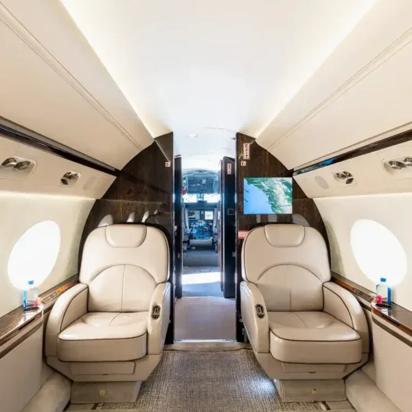 A view of the aircraft cabin interior featuring two large, light-colored leather seats facing each other, with a map displayed on a wall-mounted screen.