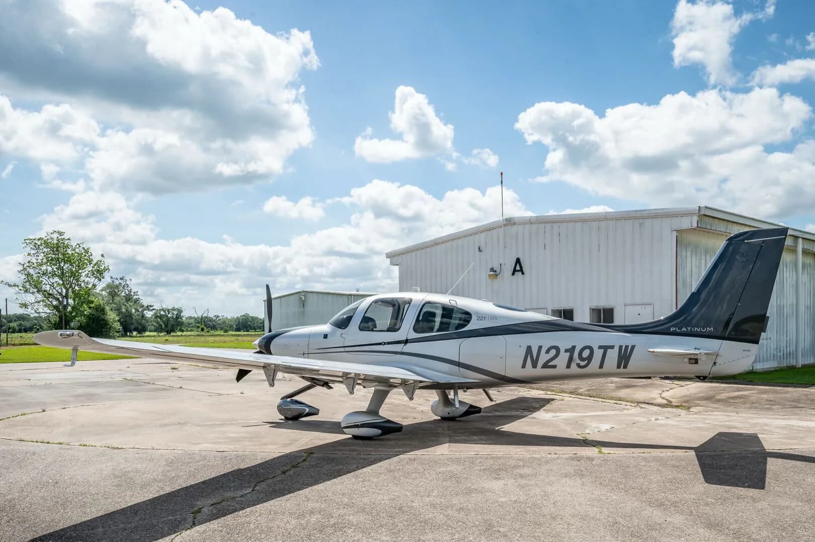 A side view of the Cirrus SR22T GTS Platinum aircraft N219TW parked on an airfield with a hangar and trees in the distance.